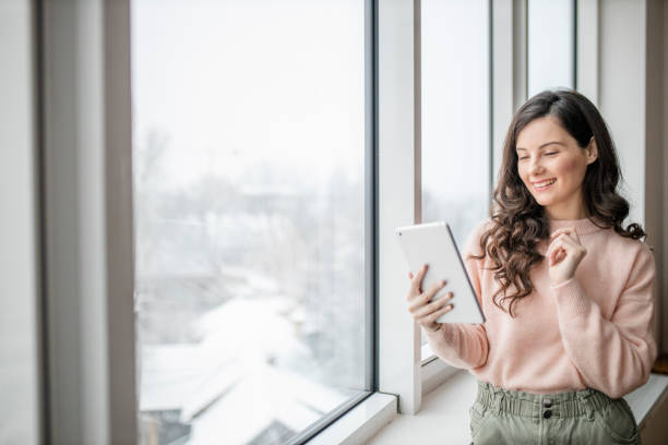 istock Beautiful young woman using her digital tablet and standing by window at home 1296882185