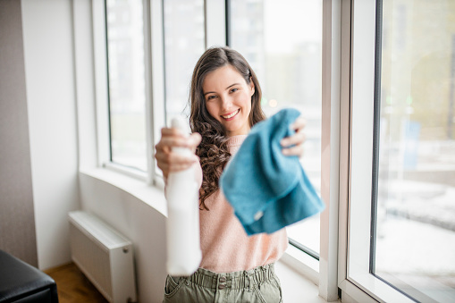Beautiful smiling young woman cleaning and wiping window with spray bottle and rag