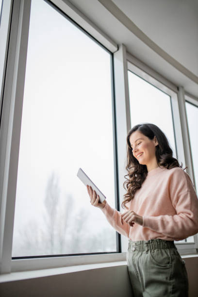 istock Beautiful young woman using her digital tablet and standing by window at home 1296886143