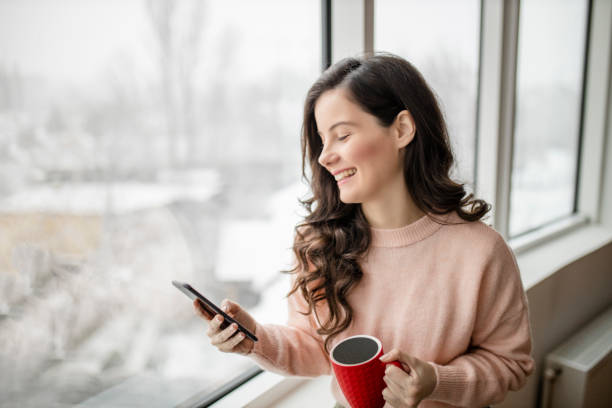 istock Woman drinking coffee, using her phone and day dreaming while looking through window 1296886469