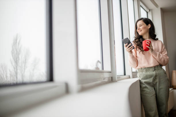 kobieta pije kawę, używając swojego telefonu i dzień marzy podczas patrzenia przez okno - reading newspaper break tea zdjęcia i obrazy z banku zdjęć