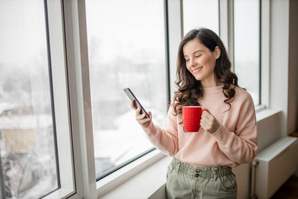 istock Woman drinking coffee, using her phone and day dreaming while looking through window 1296886806