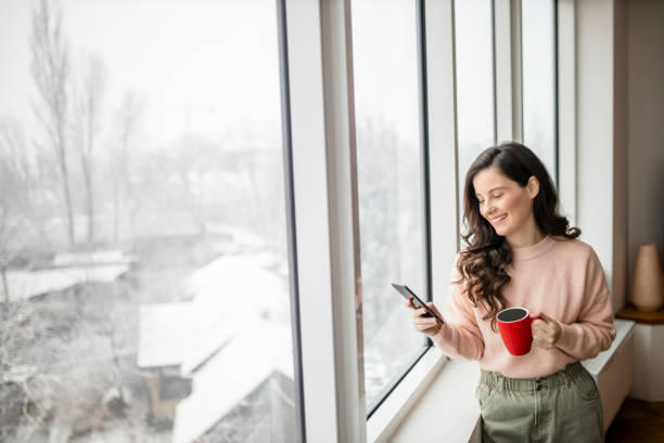istock Woman drinking coffee, using her phone and day dreaming while looking through window 1296889078