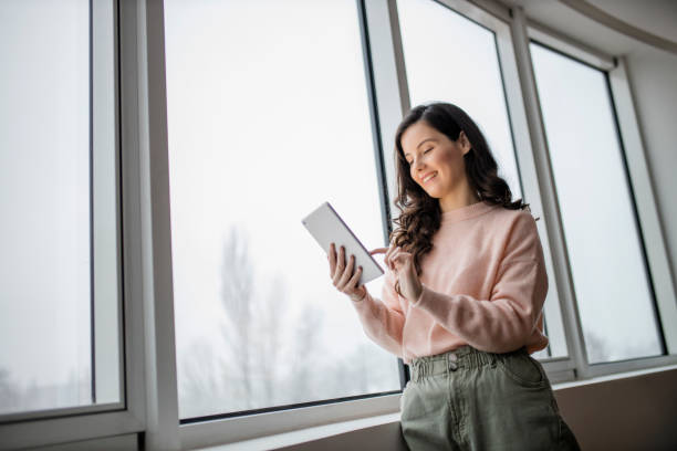 istock Beautiful young woman using her digital tablet and standing by window at home 1296889412