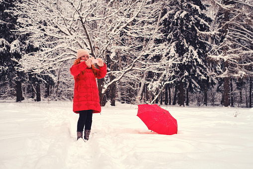 A woman with a red umbrella takes photos and videos on her phone in a winter forest. Smiling woman in red jacket with the phone in the park