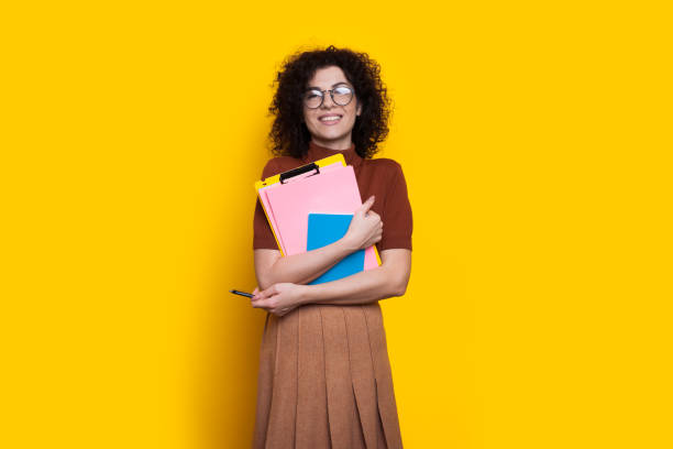 istock Lovely student with some books is smiling through glasses on a yellow studio wall 1296892479