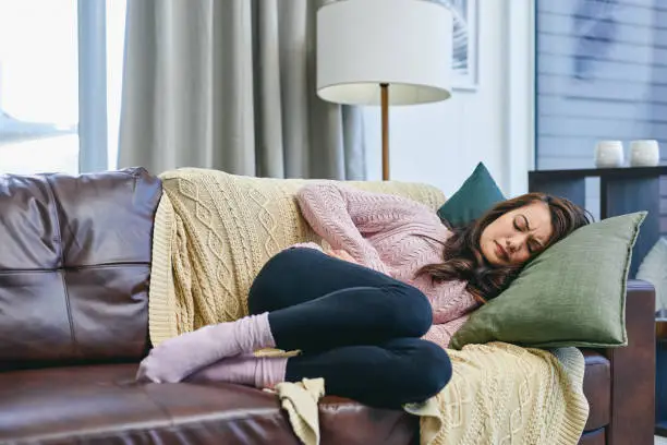 Shot of a woman looking uncomfortable while lying on the couch at home