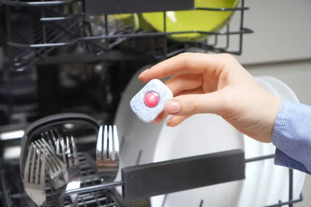 Dishwasher tablet (detergent) in female hand on the dishwasher machine backdrop
