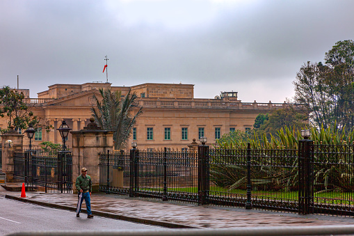 Bogota, Colombia - April 04, 2016: The Casa de Nariño as viewed from the Northeast, from Carrera Séptima in the Andean capital city. It is the official residence and the executive office of the President. It has been raining and the street is wet; the overcast sky and dark clouds indicate more rain to come. A local Colombian man is seen walking by as he goes about his afternoon work. Horizontal format.