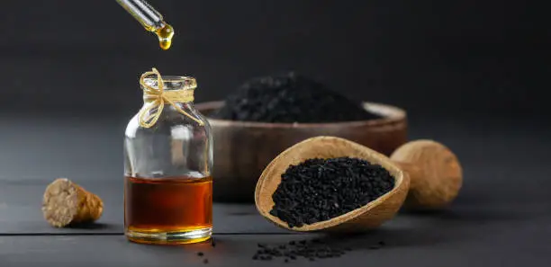 Glass bottle of black cumin seed essential oil , Nigella Sativa in scoop on black wooden background, oil dripping