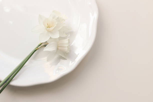 istock Styled stock photo. Spring, Easter feminine desktop scene with white narcissus, daffodils flowers on porcelain plate. Beige table background. Floral composition, web banner. Flat lay, top view. 1296905723