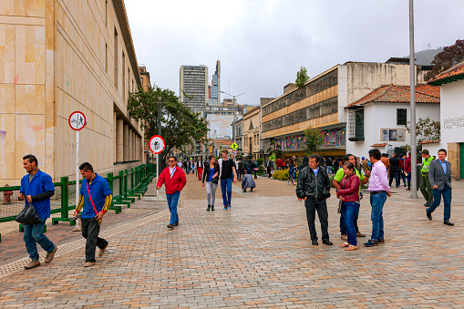 Bogota, Colombia - July 01, 2016: Carrera Septima once called Calle Real or Royal Street, at the point where it leads into Plaza Bolivar in the Andean capital city. This section of the Carrera has been closed to vehicular traffic. It is now a 'pedestrian and cycles only' zone. Image shows local Colombian people going about their daily routine. To the left are the walls of the Supreme Court of the Country. In the far background in the cloudy haze is the peak of Monserrate on the Andes Mountains. The altitude at street level is about 8,500 feet above mean sean level. People generally wear a jacket or another layer of clothing to keep warm. Photo shot in the afternoon sunlight on a rather cloudy, overcast afternoon; horizontal format.