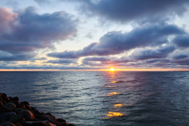 A bright orange sun sitting near coast of boulder rocks on cloudy evening sky