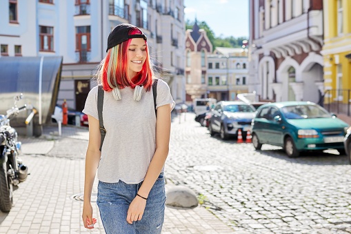 Outdoor portrait of smiling teenage student girl in black cap with backpack, fashionable female with colored dyed hair on city street, copy space. Lifestyle, youth, fashion, beauty, young people