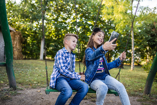 Little boy and girl are taking picture of themselves for selfie with phone while sitting on swing