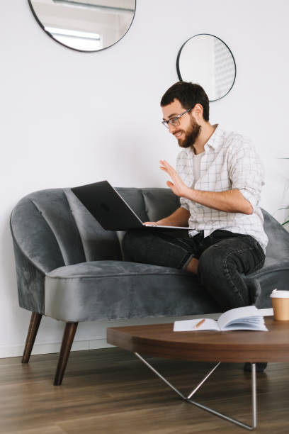 istock Smiling man sitting on a sofa, looking at laptop screen on video call 1296920195