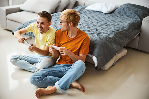 Two handsome young men sitting on the floor and smiling while using smartphones as joystick controllers