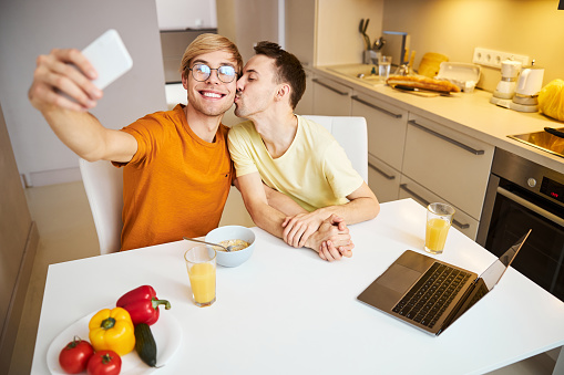 Cheerful young man taking picture with smartphone and smiling while loving boyfriend kissing his cheek