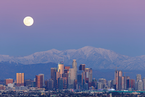 Telephoto view of a full moon rising behind the Los Angeles skyline and the San Gabriel mountains at night.
