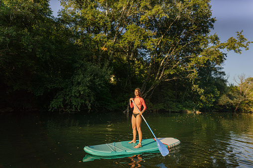 Photo of a teenage girl stand up paddling on the river; enjoying her summer vacation while being in nature and spending time outdoors.