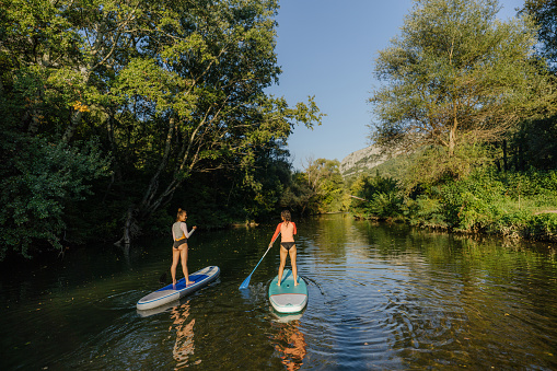 Photo of teenage girls stand up paddling on the river on a hot, sunny day; teenagers hanging out in nature and enjoying their summer vacation.