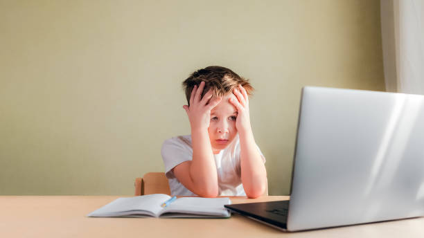 子供は机の上に座り、悲しそうにコンピュータを見て、手で頭を抱えている - learning boredom studying child ストックフォトと画像