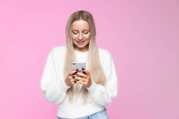 istock Cheerful young woman smiling and using mobile phone, typing sms, message, isolated over studio pink background 1296941783