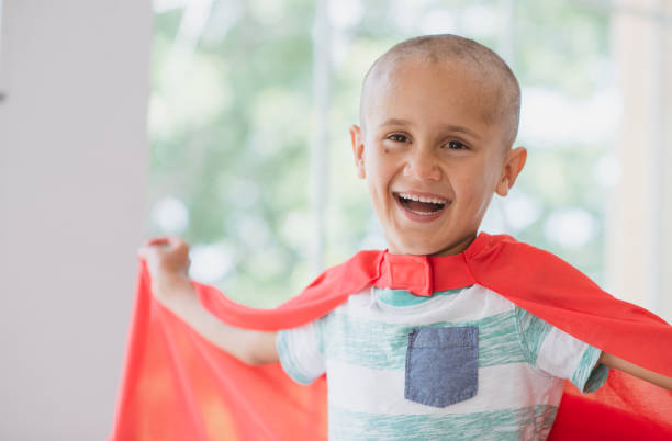 I beat cancer! A Caucasian boy around 7 years of age smiles widely at the camera as he is finally cancer free. He wears a red cape as he is pretending to be Superman. completely bald stock pictures, royalty-free photos & images