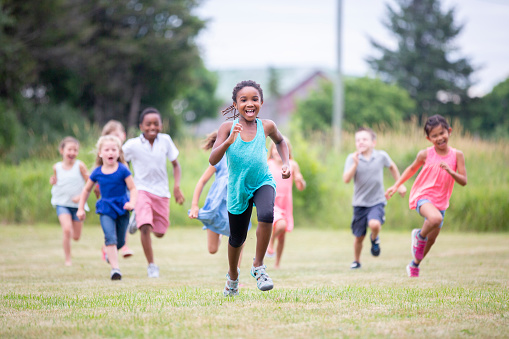 A multi ethnic group of elementary age children are running outdoors at the park. They are spending time together on a warm summer day. A girl of African ethnicity is leading the race at the front.