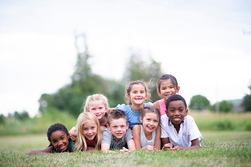 A beautiful multi ethnic group of elementary age children are all huddled together on the grass for a photo. They are all looking at the camera.