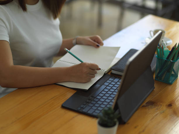 istock Female hand writing on blank notebook while working with tablet on wooden table 1296950616
