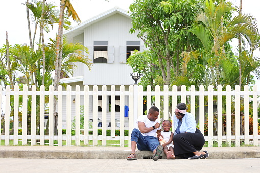 Family of African American people with young little daughter sitting in front of new house with white picket fence