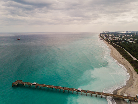 Aerial Views of the empty, blue Juno Beach seashore. Teal waves sweeping onto the clean shore. Blue skies with puffy clouds in the background.