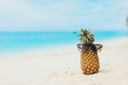 Ripe attractive pineapple in sunglasses and pink dragon fruit on the sand tropical beach against turquoise sea water, Thailand. Summer vacation concept