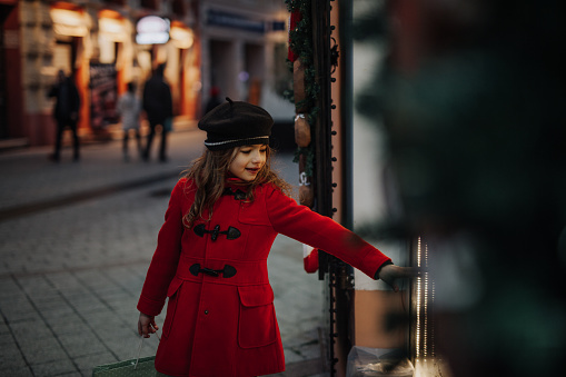Cute little girl standing in a red coat standing next to the toy window on the street.