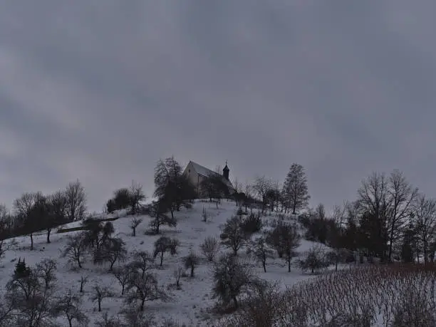 View of snow-covered hill Kapellenberg with bare trees located near Wurmlingen, Baden-Württemberg, Germany in winter season with historic chapel Sankt-Remigius-Kapelle on top on cloudy day.
