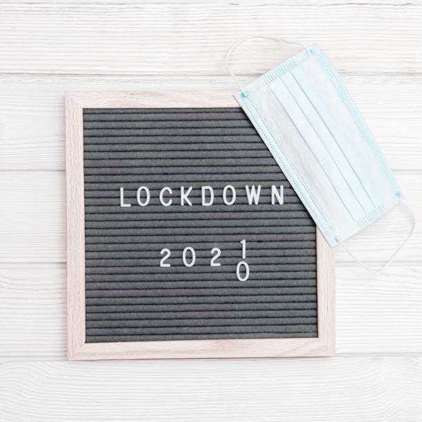 istock Flatlay letter board with message text lockdown 2021 and grey cloth protective face mask. Lock down loading concept. 1296971305