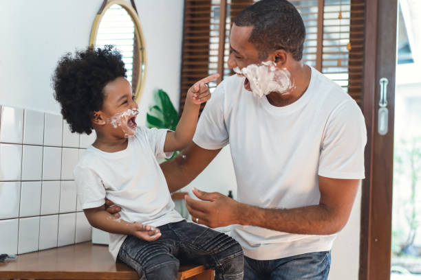 african american ojciec i synek spędzają razem czas bawiąc się śmiejąc się podczas golenia pianki na twarzach w łazience. poranna rutyna w koncepcji łazienki. - men shaving human face bathroom zdjęcia i obrazy z banku zdjęć
