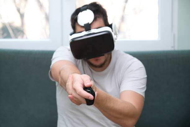 istock Young man playing a shooting video game with virtual reality headset and a joystick sitting on sofa. Having fun at home. 1296979788