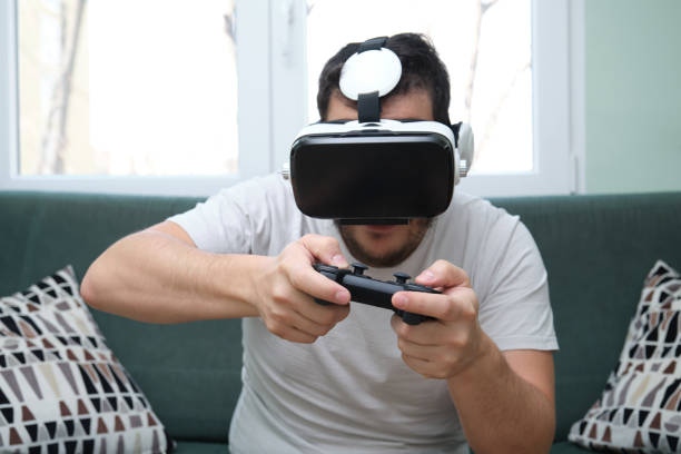istock Young man playing video games with virtual reality headset and a gamepad sitting on sofa. Having fun at home. 1296979799