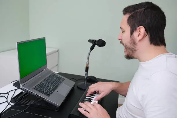 Photo of A man blogger, youtuber, influencer recording a video playing the electronic piano and singing at home. Musician, singer, piano player. Green screen on the laptop.