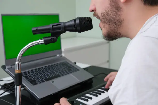 Photo of Unrecognizable man blogger, youtuber, influencer recording a video playing the electronic piano and singing at home. Musician, singer, piano player. Green screen on the laptop.