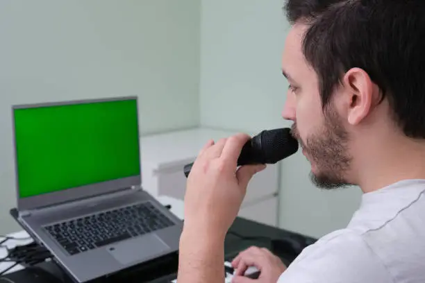 Photo of A man blogger, youtuber, influencer recording a video playing the electronic piano and singing at home. Musician, singer, piano player. Green screen on the laptop.