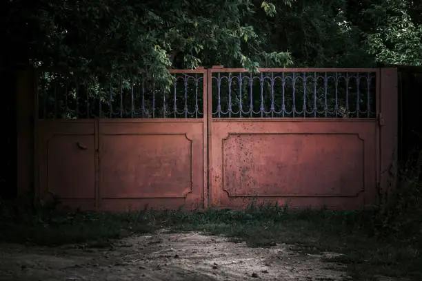 Photo of Horror concept. Iron scary rusty old closed metal gates with mystical shadows in a dark forest against the background of trees causing fear, the entrance to a gloomy cemetery in the park