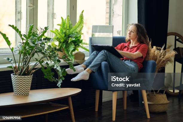 Young Female Using Laptop After Work Communicates On Internet With Customer Happy Freelance Woman Is Relaxing On Comfortable Couch And Using Laptop At Home Stock Photo - Download Image Now