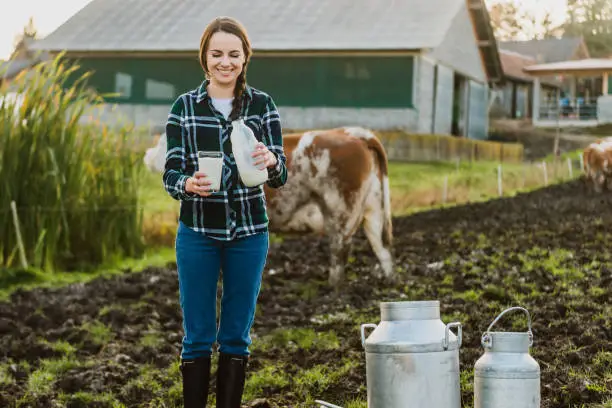 Young female farmer holding milk bottle with glass at dairy farm