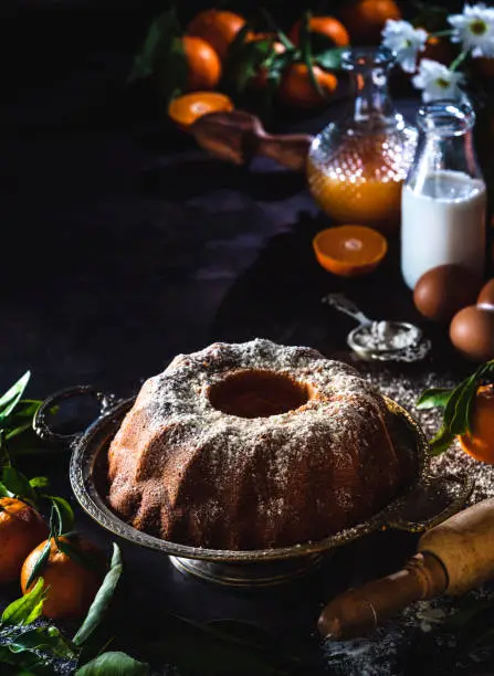 Bundt cake made of tangerine clementines baked homemade with ingredients on moody dark background