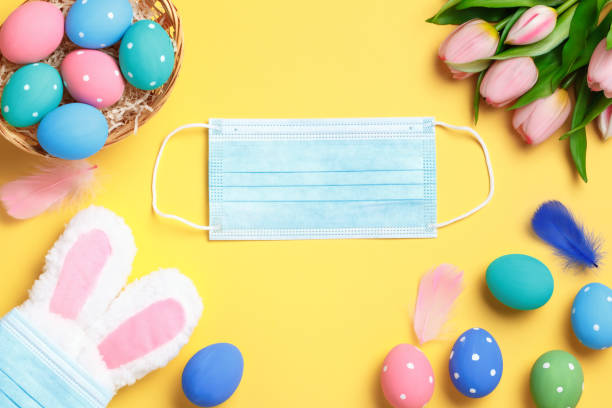 istock Happy easter. Multi-colored eggs, rabbit ears, tulips and paints on a yellow background. Flat lay greeting card. Copy the location on the medical mask. 1297012861