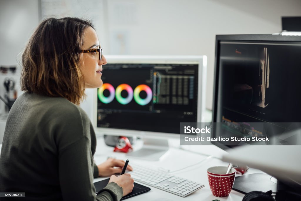 Female graphic designer at desk in the office with two displays. Profile view of Female graphic designer at the office with two displays, editing photos and video with a pen tablet. Business woman working concentrated. Freelance worker at Creative office studio. Design Professional Stock Photo