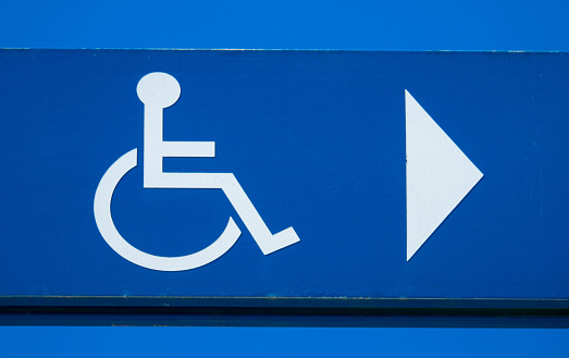 Wheelchair and arrow directional navigation icon sign post on blue sky background, handicapped helping sign in public place